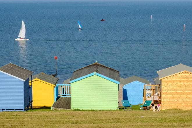 Colourful beach huts on top of a slope with a view of the sea and sail boats