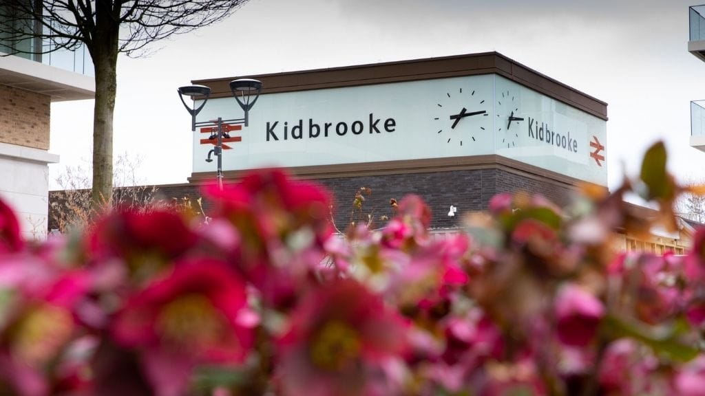 pink flowers in the foreground with Kidbrooke station in the background. Kidbrooke is written in big letters on side of the building next to a big clock face.