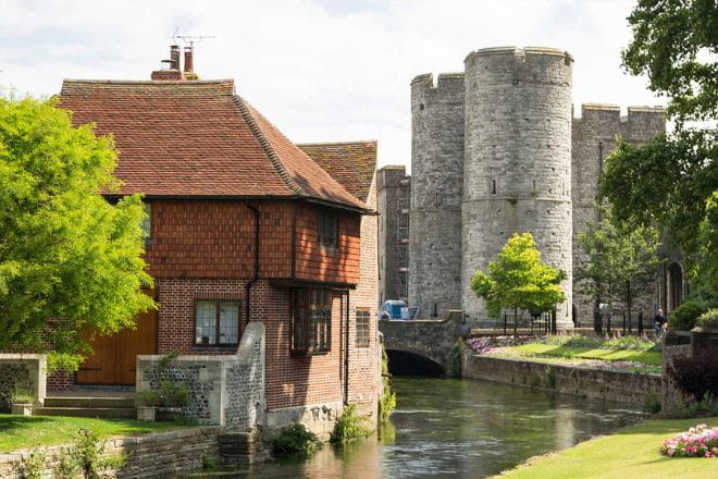 a castle with water in front of a house with Westgate, Canterbury in the background