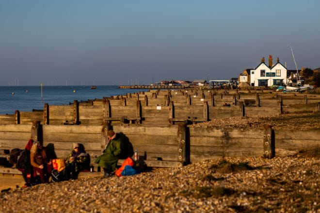 rows of wooden barriers on a beach with a pub in the distance and a family in the foreground